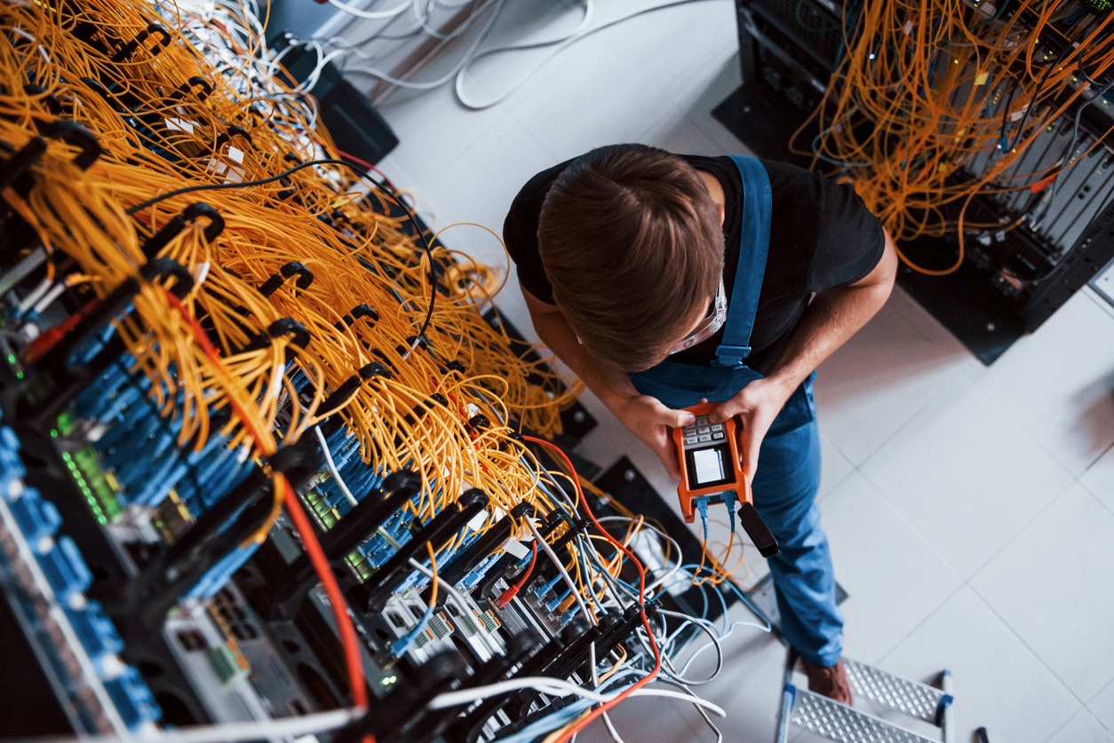 Top view of young man in uniform with measuring device that works with internet equipment and wires in server room